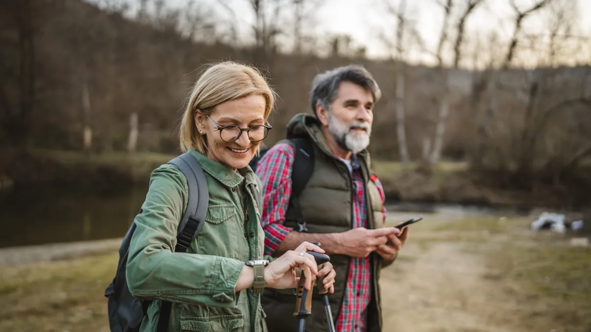 Couple hiking by a river — she checks her smartwatch while he holds a phone